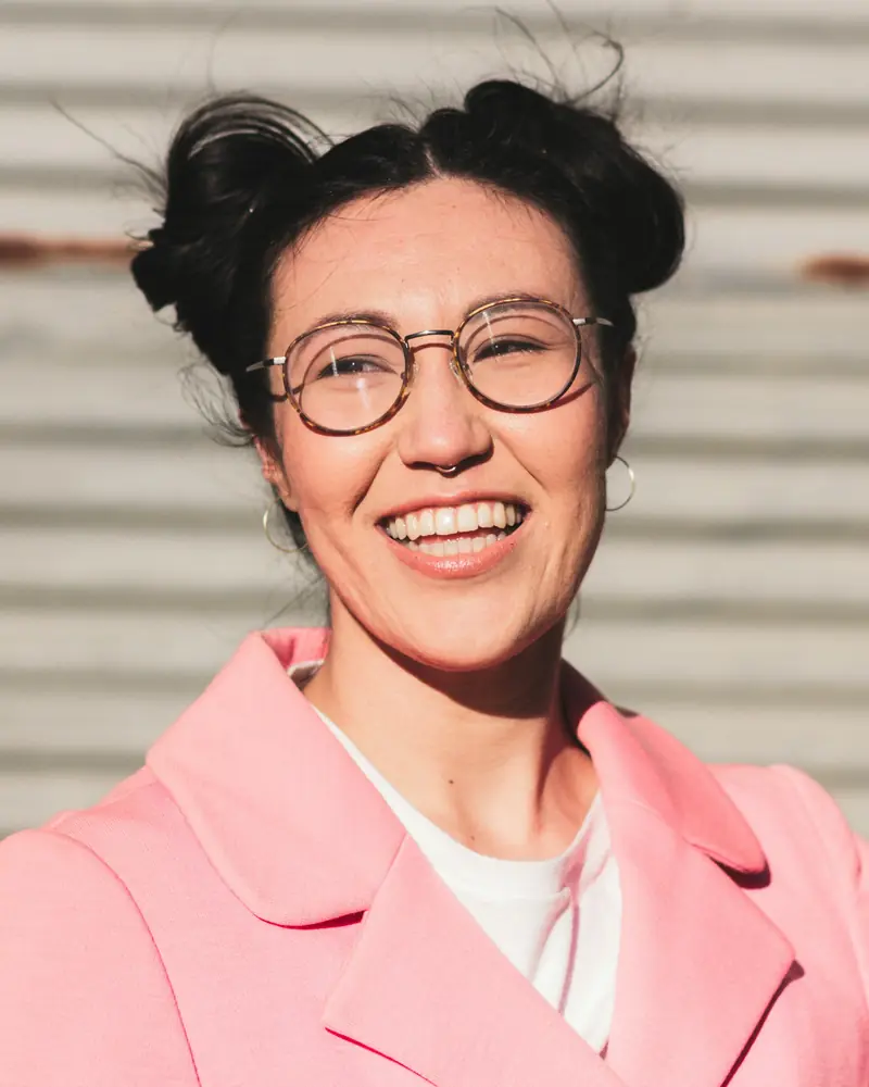 A woman in round glasses and a pink blazer laughing in bright flash light