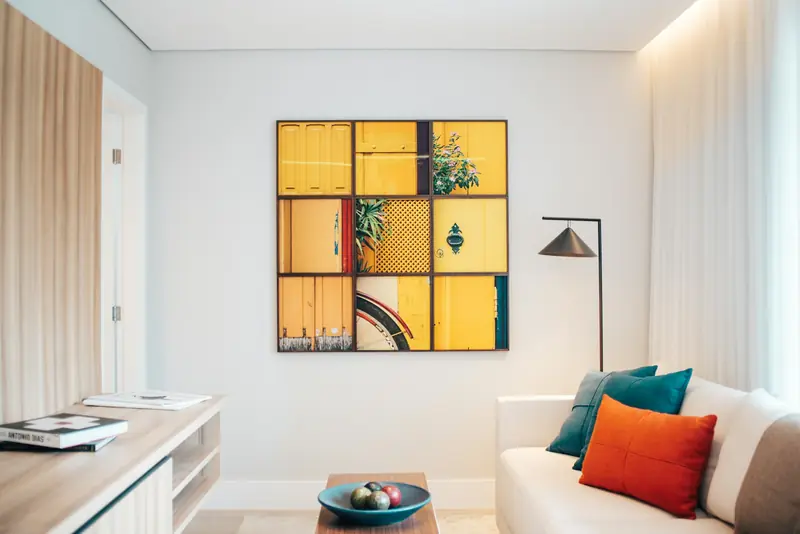 A white wall with a yellow-paned window artwork above orange cushions
